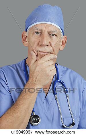 Stock Photo - Portrait of senior male surgeon with hand on chin over gray background. Fotosearch
