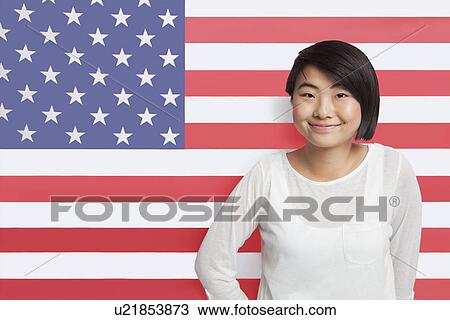 Stock Image - Portrait of young Asian woman smiling against American flag. Fotosearch