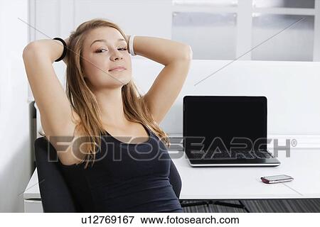 Stock Photo - Portrait of young businesswoman with hands behind head sitting in front of desk in office. Fotosearch