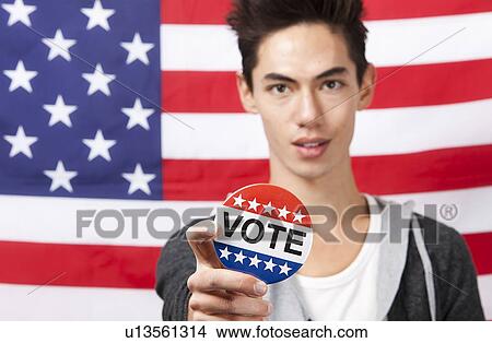 Picture - Portrait of young man displaying vote badge against American flag. Fotosearch