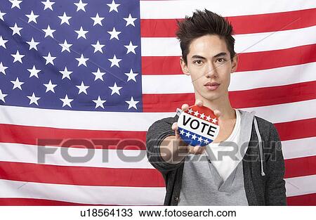 Stock Image - Portrait of young man showing vote badge standing against American flag. Fotosearch