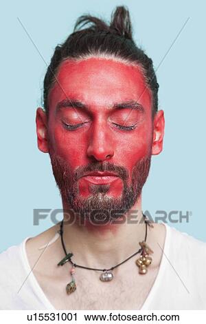Stock Image - Portrait of young man with eyes closed and red painted face against blue background. Fotosearch