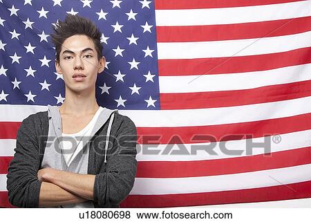 Stock Photo - Portrait of young mixed race man with arms crossed standing against American flag. Fotosearch