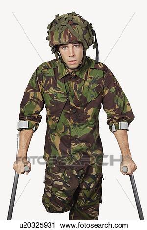 Stock Image - Portrait of young soldier holding crutches against gray background. Fotosearch
