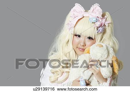 Stock Photograph - Portrait of young woman dressed as a doll holding soft toy over gray background. Fotosearch