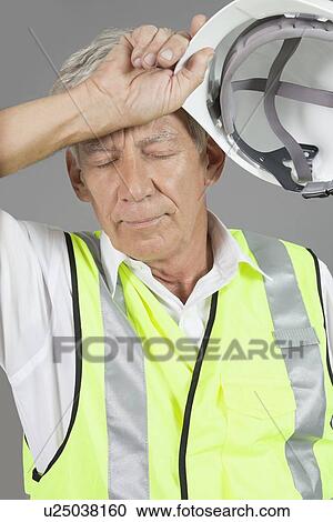 Stock Image - Senior construction worker wiping his brow over gray background. Fotosearch