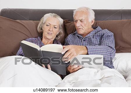 Stock Photo - Senior couple with digital tablet reading book while lying on bed in room. Fotosearch