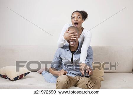 Surprised young woman covering man's eyes while watching TV View Large Photo Image Stock Image - Surprised young woman covering man's eyes while watching TV. Fotosearch