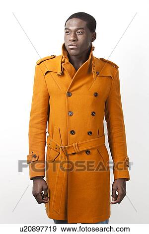 Stock Photo - Young African American man in orange trench coat standing against white background. Fotosearch