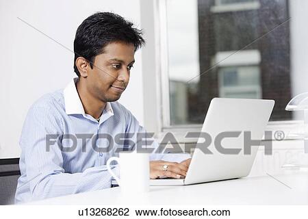 Young businessman using laptop at desk in office View Large Photo Image Stock Image - Young businessman using laptop at desk in office. Fotosearch