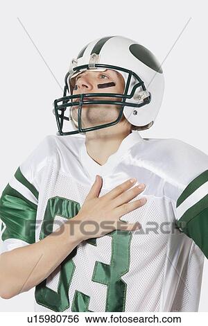 Stock Photograph - Young man in football uniform with hand on chest against gray background. Fotosearch