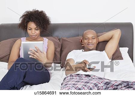Stock Image - Young man watching TV while woman using tablet PC in bed. Fotosearch