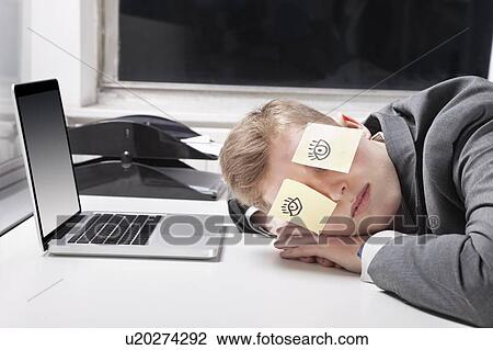 Stock Image - Businessman sleeping with sticky notes on eyes at desk in office. Fotosearch