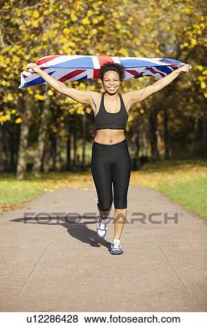 Happy young female jogger holding up British flag on pathway in park View Large Photo Image Stock Photo - Happy young female jogger holding up British flag on pathway in park. Fotosearch