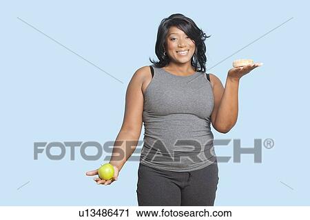 Overweight mixed race woman holding green apple and donut over blue background View Large Photo Image Stock Image - Overweight mixed race woman holding green apple and donut over blue background. Fotosearch