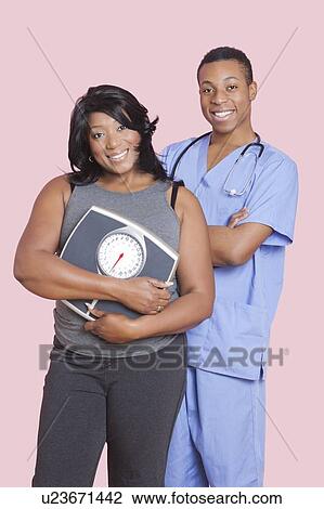 Stock Image - Overweight mixed race woman holding scales over pink background. Fotosearch