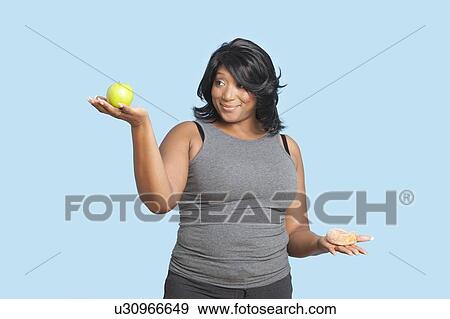 Stock Photo - Overweight mixed race woman holding green apple and donut over blue background. Fotosearch