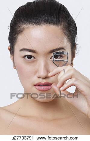 Stock Image - Portrait of beautiful young woman using eyelash curler against white background. Fotosearch