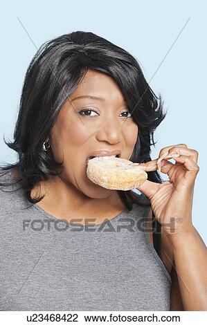 Stock Image - Portrait of overweight mixed race woman eating donut over blue background. Fotosearch