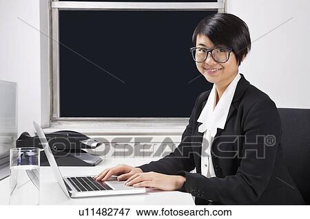 Stock Photo - Portrait of smiling young businesswoman using laptop at desk in office. Fotosearch