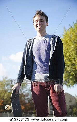 Stock Image - Portrait of young man with skateboard smiling against sky. Fotosearch