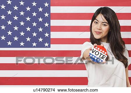 Picture - Portrait of young woman holding out vote badge against American flag. Fotosearch