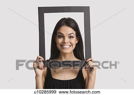 Stock Photo - Portrait of young woman smiling through frame over white background. Fotosearch