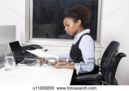 Stock Image - Side view of young businesswoman with document using laptop at desk in office. Fotosearch