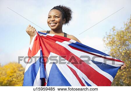 Stock Photography - Smiling young woman holding British flag against trees and sky. Fotosearch