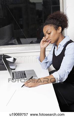Young businesswoman using cell phone in front of laptop at desk in office View Large Photo Image Stock Photo - Young businesswoman using cell phone in front of laptop at desk in office. Fotosearch