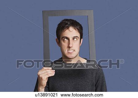Stock Image - Young man making funny faces through frame over blue background. Fotosearch