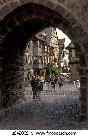 Stock Photography -  Alsace, France, Europe, Riquewhir, Haut Rhin, People walking on the cobbled streets in the village of Riquewhir. Through the arch half-timbered houses (Alsatian-style buildings) can be seen.. Fotosearch