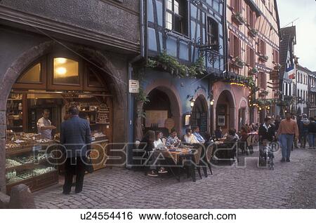 Stock Photograph -  Alsace, outdoor cafe, France, Rikewhir, Europe, People sitting at an outdoor cafe watching tourists stroll down the narrow cobble streets in the picturesque village of Rikewhir with its half-timbered houses, a wine growing village of Alsace, France. . Fotosearch