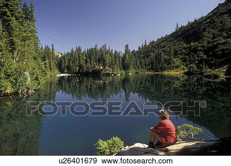 Stock Photo -  Cascades, Mt. Rainier, Mount Rainier National Park, lake, Cascade Range, Washington, A woman hiker sits on a rock looking at the reflection in the calm Snow Lake in the Cascade Mountain Range in Mt. Rainier Nat'l Park in the state of Washington.. Fotosearch