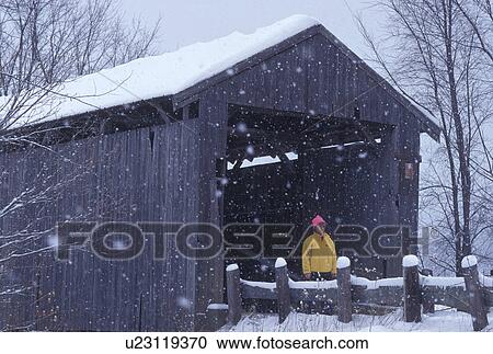 Stock Image -  covered bridge, winter, Vermont, A woman walks across the Scribner or Mudget Covered Bridge on a snowy wintry day in Johnson in Lamoille County in the state of Vermont.. Fotosearch