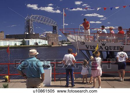 Stock Photography -  Duluth, Minnesota, Passengers on The Vista Fleet/Duluth-Superior Excursion tourboat with people watching from the waterfront along the St. Louis River with the Aerial Lift Bridge in the background in the city of Duluth in the state of Minnesota . Fotosearch