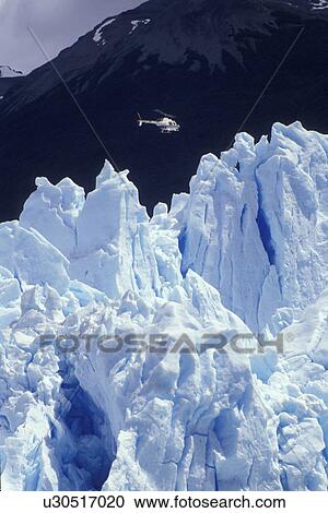 Stock Image -  glaciers, Patagonia, Argentina, Andes, A helicopter looks tiny flying over the massive Moreno Glacier which rises 60 meters above lake level in Parque Nacional de los Glaciares (National Park) in Patagonia. . Fotosearch