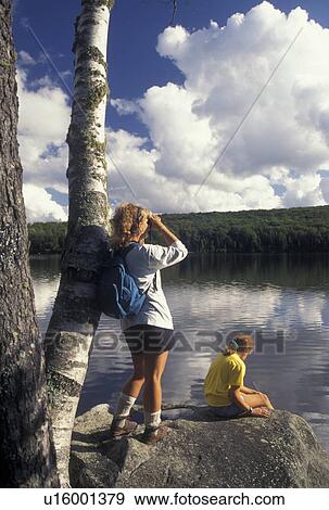 Stock Photo -  hiking, Vermont, Mother and daughter taking a break after hiking around Kettle Pond in Groton State Forest in Caledonia County in the state of Vermont. Mother standing on boulder leaning against a birch tree looking through binoculars, daughter sitting o. Fotosearch