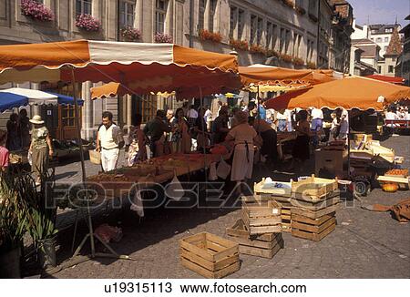 Stock Image -  market, Switzerland, Lausanne, Vaud, Europe, People shopping at a produce stand with orange umbrellas at a street market at Place de la Palud in Lausanne in the Canton of Vaud.. Fotosearch