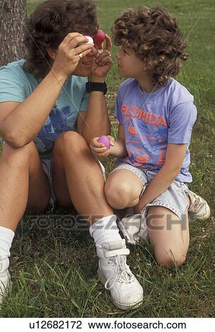 Stock Image -  mother, child, easter, eggs, 6 year old daughter watches her mother make a funny face putting two colored Easter Eggs up to her eyes while sitting on the lawn in a park on a warm spring day.. Fotosearch
