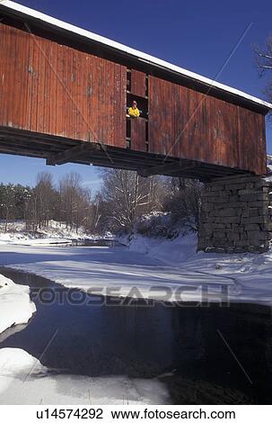 Stock Image -  red covered bridge, winter, Vermont, A woman stands in the window of The Slaughter House Covered Bridge which crosses over the partially frozen water of the Dog River on a winter day in Northfield Falls in Washington County in the state of Vermont.. Fotosearch