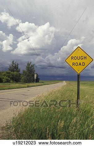 Stock Image -  storm clouds, road, rough road, North Dakota, A yellow rough road sign is posted next to a isolated road with storm clouds in the distance in the state of North Dakota.. Fotosearch