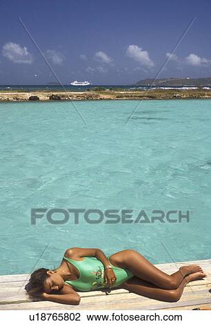 Stock Image -  sunbathing, Caribbean, Union Island, Grenadine Islands, Caribbean Islands, Woman in green bathing suit sunbathing on a dock next to the green waters of the Caribbean Sea on Union Island in the St. Vincent Grenadines (a British Commonwealth member).. Fotosearch
