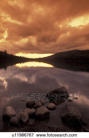 Stock Photo -  sunset, lake, Vermont, Orange clouds reflect in the calm waters of Kettle Pond at sunset with boulders in the foreground in Groton State Forest in Caledonia County in the state of Vermont. . Fotosearch