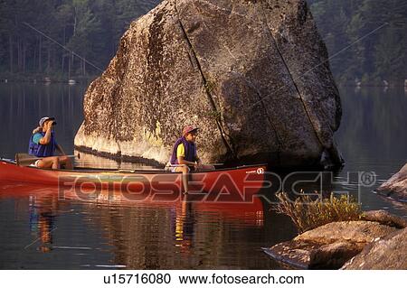Stock Image -  Vermont, canoe, canoeing, Mother and daughter sitting together in canoe on Kettle Pond next to a huge boulder in Groton State Forest. Daughter relaxing with feet in water. Mother looking through binoculars. Mad River Canoe Products.. Fotosearch
