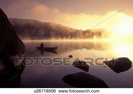 Stock Photograph -  Vermont, canoe, canoeing, Woman paddles canoe in the early morning [fog, mist] on Kettle Pond at[sunrise, sunset] in Groton State Forest. Mad River Canoe Products.. Fotosearch