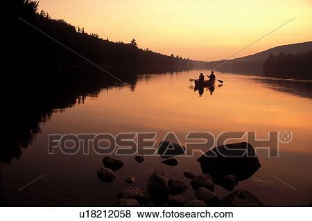 Stock Photo -  Vermont, canoeing, Two people paddling a canoe over the quiet calm waters of Kettle Pond at[sunrise, sunset] in Groton State Forest. Mad River Canoe Products.. Fotosearch