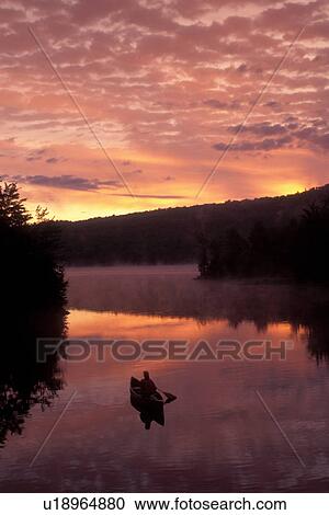 Stock Image -  Vermont, canoeing, Woman paddles a canoe over the quiet calm waters of the Green River Reservoir at [sunrise, sunset]. Mad River Canoe Products.. Fotosearch
