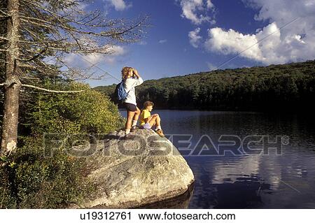 Stock Image -  Vermont, Mother and daughter [sitting, standing] on a boulder taking a break from hiking on Kettle Pond in Groton State Forest. Woman looking through binoculars . Fotosearch