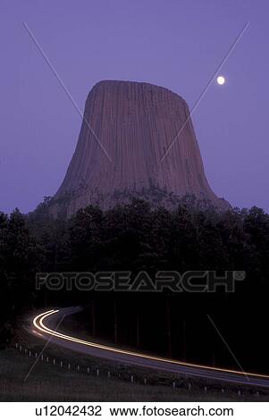 Stock Image -  Wyoming, Full moon rises over Devils Tower National Monument at night. Car lights streak down the road away from the tower.. Fotosearch